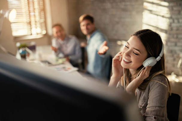 Happy female entrepreneur wearing headphones and enjoying in music with her eyes closed in the office. Her colleagues are in the background.  - Foto, Imagen