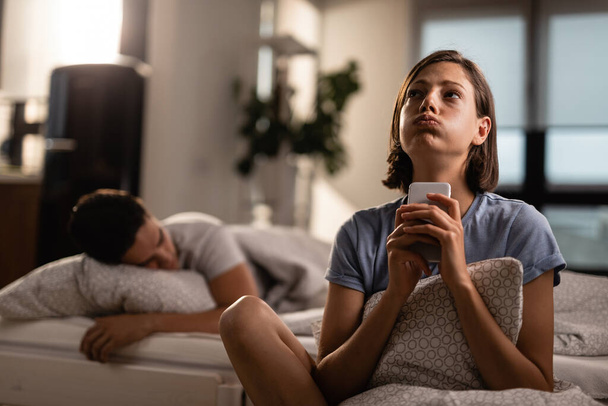 Сумнівна жінка використовує мобільний телефон і думає про щось, поки її хлопець спить на ліжку позаду неї
. - Фото, зображення