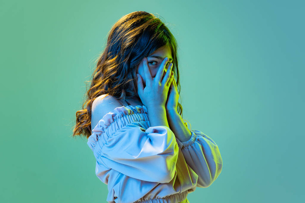 Shyness. Beautiful cute little girl, kid closes her face with hands isolated over cyan color background in white neon light. Concept of children emotions, fashion, beauty, back to school and ad - Foto, Imagem