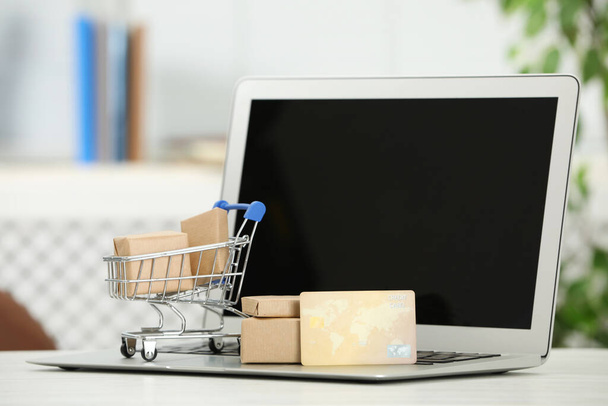Online payment concept. Small shopping cart with bank card, boxes and laptop on white table - Fotoğraf, Görsel