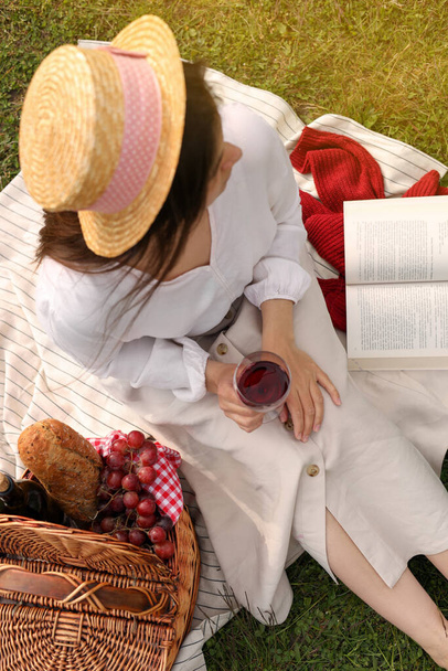Woman with glass of wine, book and picnic basket on green grass, above view - Fotó, kép