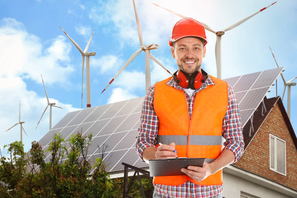 Industrial engineer in uniform and view of wind energy turbines near house with installed solar panels on roof - Photo, Image