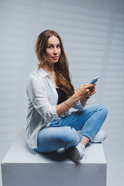 Młoda kobieta z długimi brunetkami, ubrana w białą koszulę i dżinsy, rozmawiająca przez telefon. Kobieta w dżinsach i biała koszula z telefonem komórkowym. Białe tło - Zdjęcie, obraz