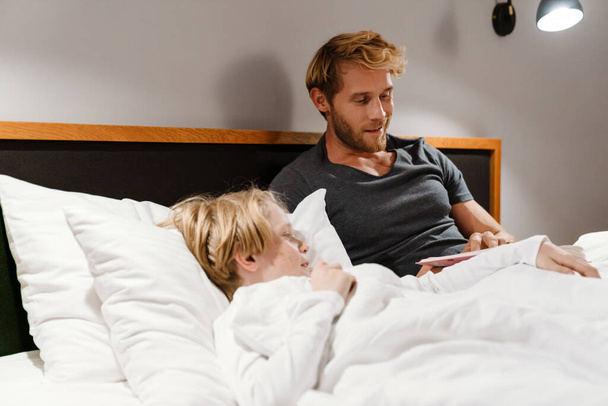 White man reading book to his son while lying in bed at home - Φωτογραφία, εικόνα