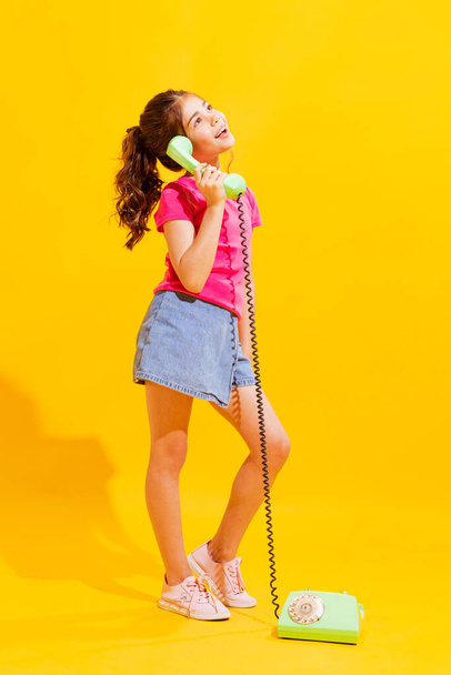 Calling. Emotional little girl, kid in casual style clothes using retro phone isolated over bright yellow background. Concept of kids emotions, fashion, beauty, back to school and ad - Photo, Image