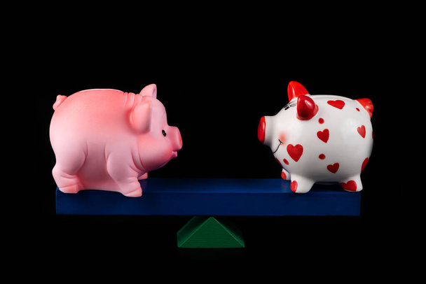 Two piggy banks on a balancing board on a black background - Zdjęcie, obraz