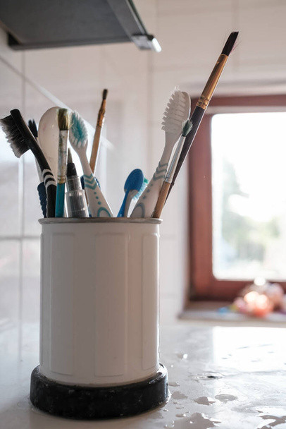 Nasse Bürsten und Zahnbürsten in einem Metallhalter, auf dem Küchentisch, im Tageslicht aus dem Fenster. Nahaufnahme. - Foto, Bild