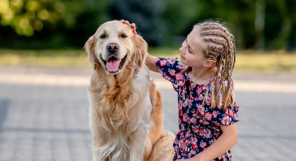Preteen girl with golden retriever dog sitting in park in beautiful summer day. Adorable female child kid hugging purebred doggy pet outdoors portrait - Φωτογραφία, εικόνα