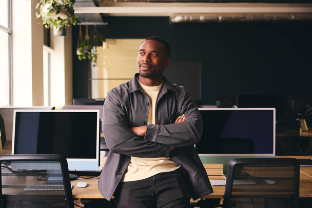 Улыбающийся молодой чернокожий мужчина стоит в офисе и смотрит в сторону от камеры с скрещенными руками - Фото, изображение