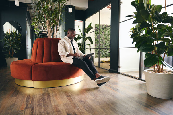 Tiro de comprimento total de homem jovem casualmente vestido trabalhando no laptop no escritório moderno em plano aberto sentado no sofá circular laranja - Foto, Imagem