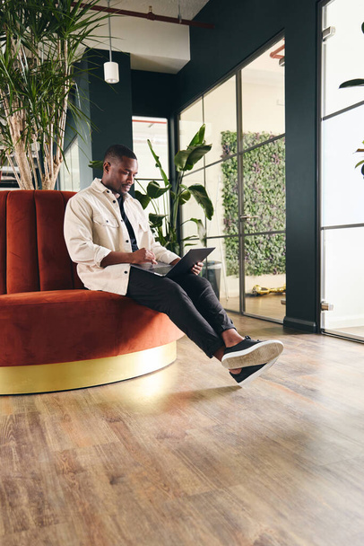 Täyspitkä laukaus rennosti pukeutunut nuori mies työskentelee kannettavan modernin avoimen suunnitelman toimisto istuu oranssi pyöreä sohva muotokuva - Valokuva, kuva