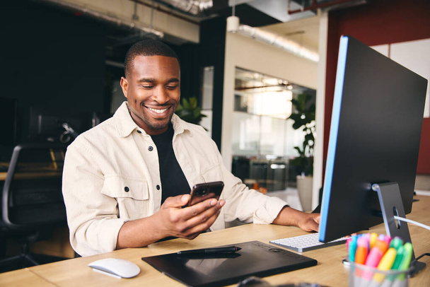 Jovem preto masculino publicidade marketing ou design criativo no escritório moderno sentado na mesa usando telefone móvel - Foto, Imagem