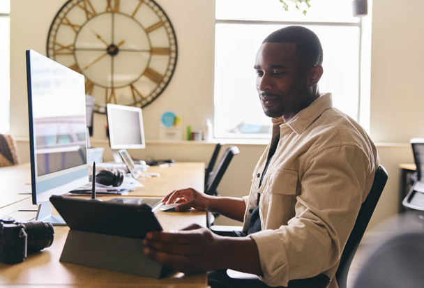 Молодой креативный черный человек, работающий за компьютером в современном офисе за столом на планшетном компьютере улыбается - Фото, изображение