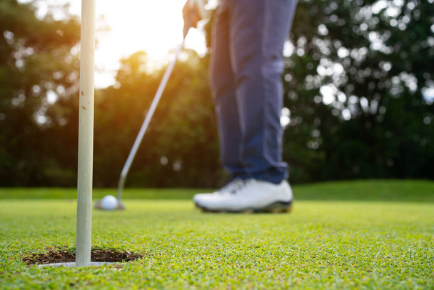 Гольфер кладет мяч на зеленый гольф, линзы вспыхивают на закате вечером. Размытый гольфист играет в гольф на красивом поле для гольфа на вечернем поле для гольфа с солнечным светом.  - Фото, изображение