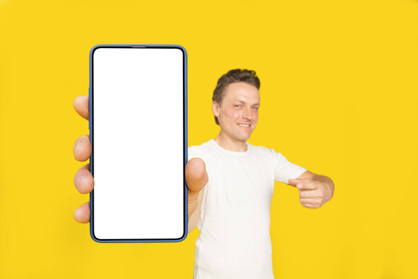 Huge smartphone in hand of handsome blond man pointing at white empty screen, wearing white t-shirt and jeans isolated on yellow background. Mobile app advertisement, great offer. - Photo, image