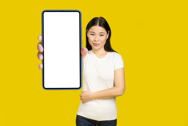 Showing phone white screen middle aged asian woman 40s holding smartphone mobile app advertisement and excited smile on camera isolated on yellow background. Product placement.  - Photo, Image