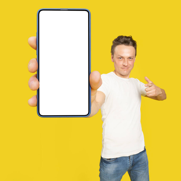 Handsome man holding huge smartphone with white screen pointing at white empty screen, wearing white t-shirt and jeans isolated on yellow background. Mobile app advertisement, great offer. - Foto, Imagem