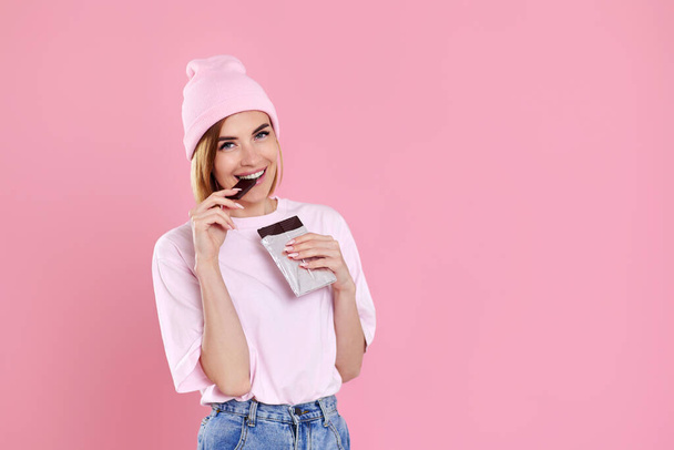young happy woman in casual t-shirt biting sweet chocolate bar and looking at camera isolated on pastel pink background - Zdjęcie, obraz