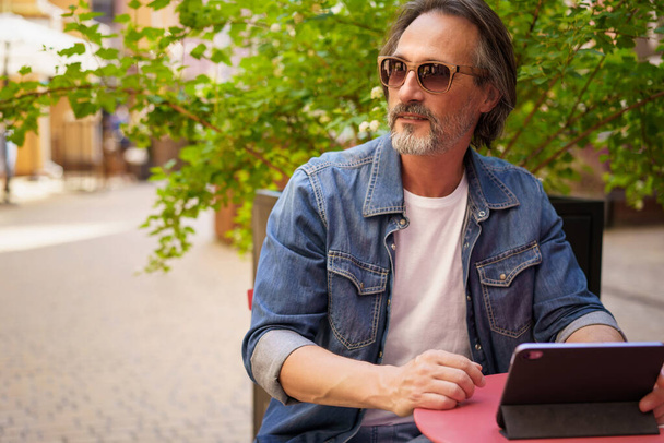 Handsome mature freelancer man enjoying coffee outdoors while having group chat. Middle aged man looking away sitting at cafe or street restaurant while working outdoors using digital tablet.  - Fotografie, Obrázek