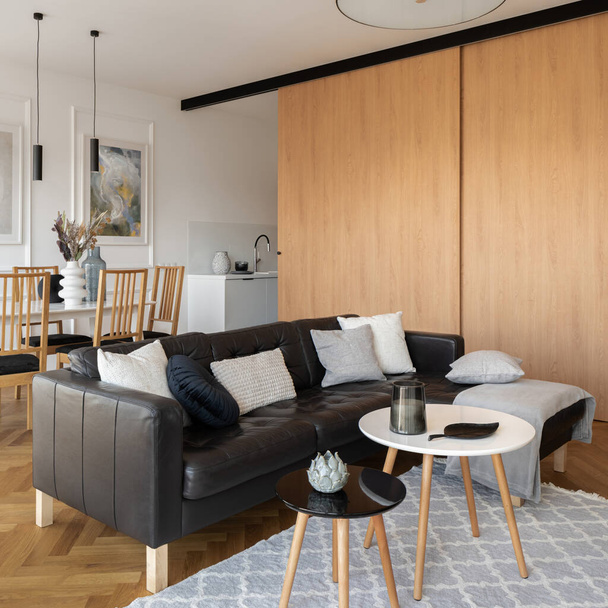 Leather black, corner sofa with pillows and simple coffee tables in living room with wooden sliding doors and kitchen open to dining table with wooden chairs - Fotó, kép