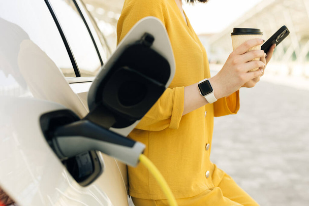 Unrecognizable woman using smart phone and waiting power supply connect to electric vehicles for charging the battery in car. Plug charging an Electric car from charging station. - Zdjęcie, obraz