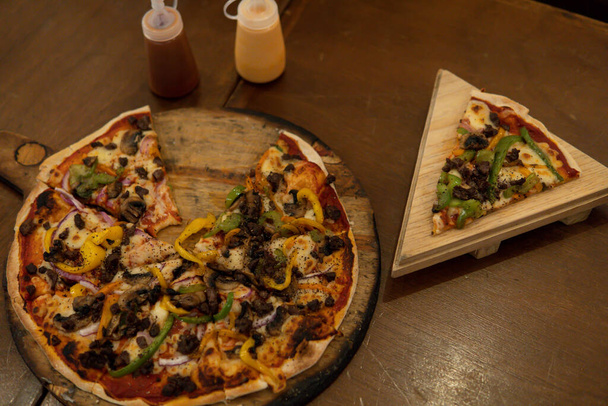 View from above of a wood oven pizza with mushrooms, meat, and green and yellow peppers. Piza with a slice cut and served on a wooden plate. Wood background. - Φωτογραφία, εικόνα