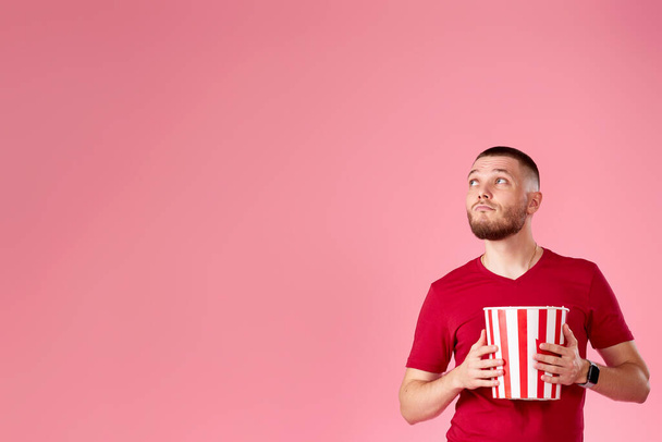 Young caucasian man eating popcorn and looking at the side on pink background. copy space - Fotoğraf, Görsel
