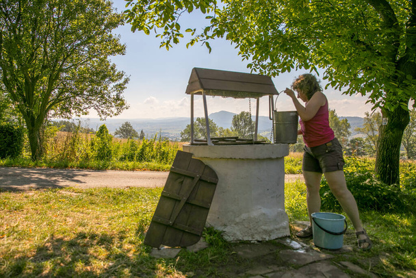 Žena bere vodu ze starého kamene, studny s větrem a klikou v polské vesnici. V pozadí pohled na kopec Cergowa v Nízkých Beskydech, Beskid Niski, Polsko - Fotografie, Obrázek