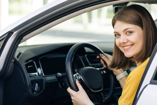Attractive young business woman looking over her shoulder while driving a car. Driver smiles at camera. Happy millennial woman taxi driver sitting in car holding to steering wheel - Foto, Imagen