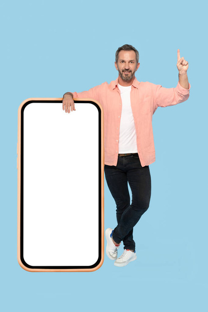 Gesturing attention pointing finger up middle aged grey haired man leaned on huge, giant smartphone with white screen happy smiling on camera wearing peach shirt isolated on blue background. - Photo, Image