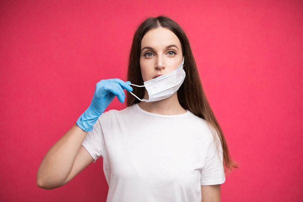 A fiatal nő orvosi maszkot visel, védekező sebészeti steril kesztyűben a karján, rózsaszín alapon izolálva, védve a koronavírus ellen. Kiváló minőségű fénykép - Fotó, kép