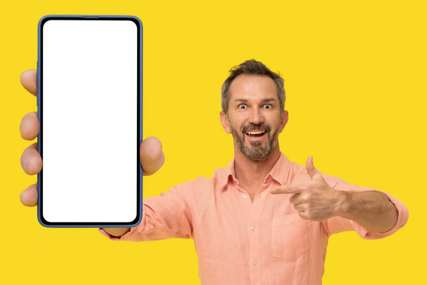 Przystojny, siwy mężczyzna w średnim wieku, wskazujący na smartfona w ręku, zadziwiający uśmiech patrząc na aparat w brzoskwiniowej koszuli odizolowanej na żółto. Dojrzałe fit człowiek z telefon aplikacja reklama.  - Zdjęcie, obraz