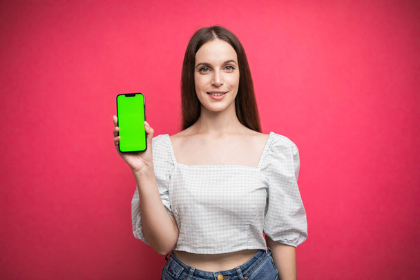 Mutlu kadın ayakta duruyor ve telefonu pembe arka planda tutuyor. Yeşil ekran telefon ekranı. Yüksek kalite fotoğraf - Fotoğraf, Görsel