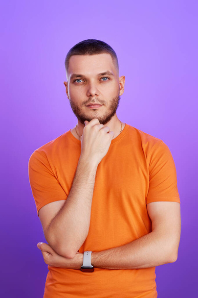 young handsome thoughtful man holding arms folded, touching chin with hand on purple background - Фото, изображение