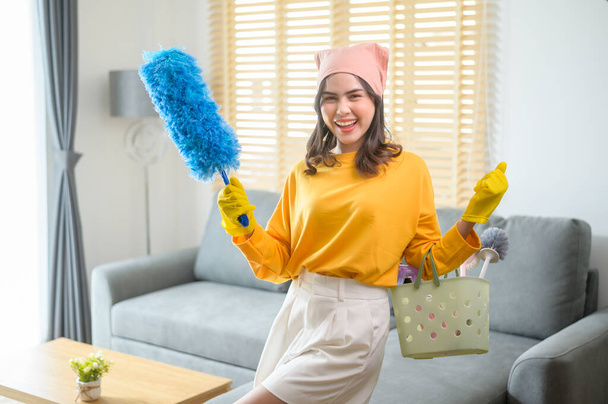 A Young happy woman wearing yellow gloves  and holding a basket of cleaning supplies in living room. - Photo, image