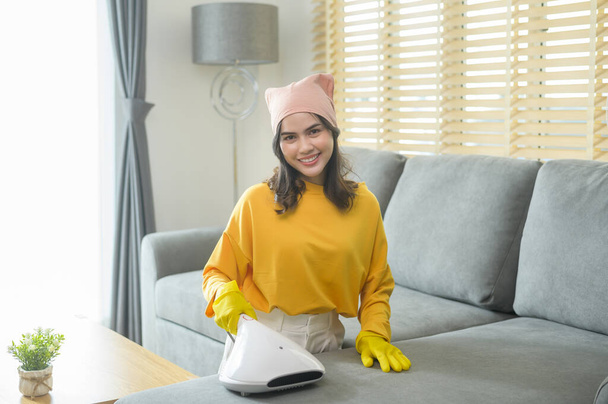 Young happy woman wearing yellow gloves  and vacuum Cleaning a sofa in living room. - Fotoğraf, Görsel