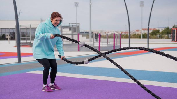 Caucasian woman in a mint sweatshirt is training with battle ropes at the sports ground - Foto, Bild