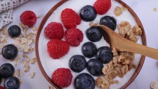 Serving a portion of yogurt with blueberry raspberry and oat flakes in a wooden bowl ready to be served. Healthy food for dieting concept. Fruit muesli with yogurt. Dolly shot 4k - Séquence, vidéo