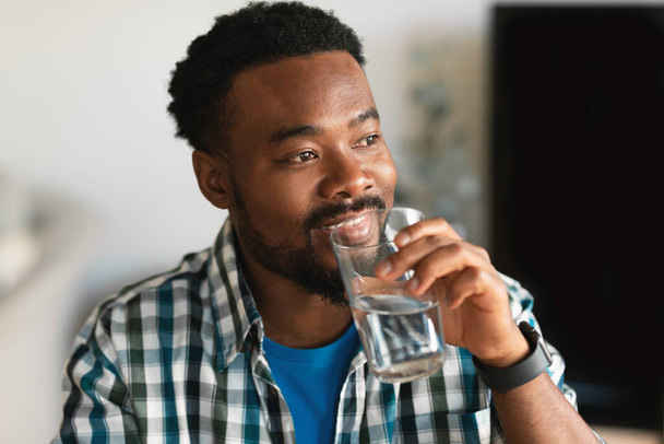 Happy Millennial African American Man Drinking Water Holding Glass Looking Aside Hydrating Sitting At Home. Healthy Lifestyle, Liquid And Hydration Concept - Foto, afbeelding