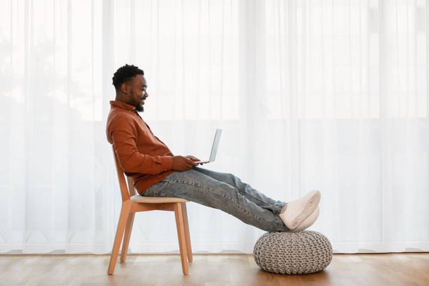 Веселый афроамериканец, сидящий в кресле возле окна дома. Человек, работающий в Интернете и удаленно подключающийся к сети. Независимая карьера. Шот с видом сбоку - Фото, изображение