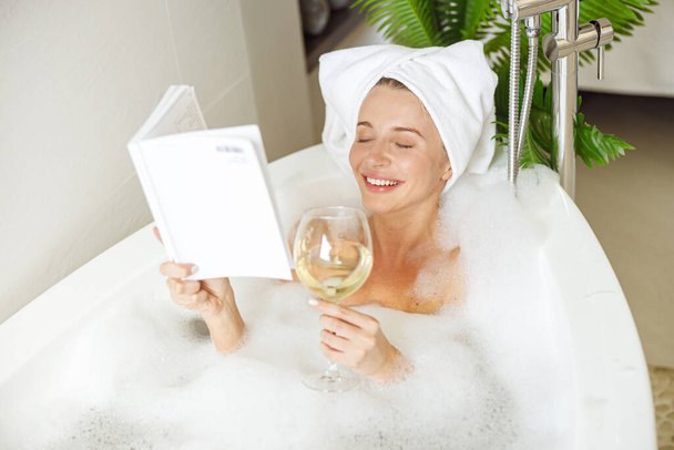 Güzel, gülümseyen genç bir kadın köpükle banyoda uzanıyor, kitap okuyor ve şarap içiyor. Sakin ol.. - Fotoğraf, Görsel
