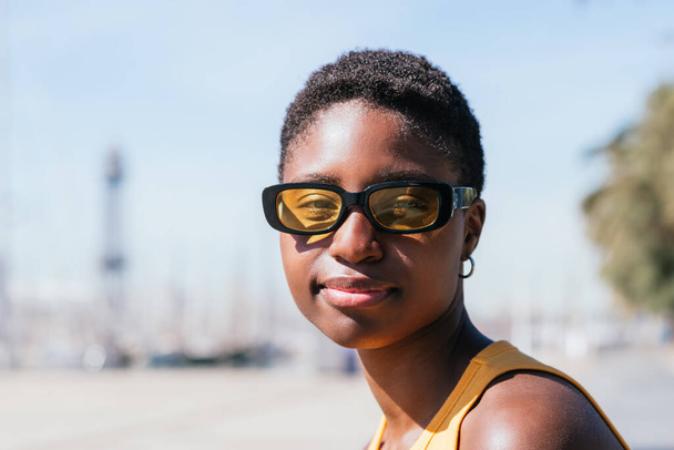 retrato horizontal de una atractiva mujer afroamericana mirando a la cámara. Lleva gafas de sol y una camiseta amarilla. - Foto, Imagen