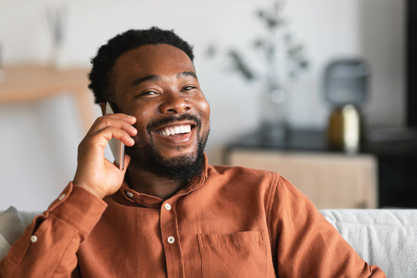 Cheerful African American Male Talking On Phone And Smiling Communicating Looking At Camera Sitting On Couch At Home. Shot Of Man Speaking By Cellphone. Mobile Communication - Foto, imagen