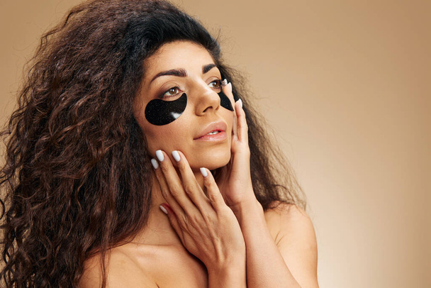 Iloinen kaunis latinalaisamerikkalainen nainen, jolla on mustia hydrogeelilaastareita silmäkosketuksen alla, näyttää syrjään poseeraten pastellin beigen taustan yli. Kosmeettinen tuote mainos Natural kauneus käsite Studio muotokuva - Valokuva, kuva