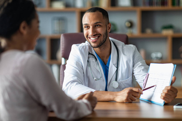 Cheerful young arabic general practitioner showing black lady patient treatment plan, african american woman attending therapist at newest private clinic, modern healthcare concept - Photo, Image