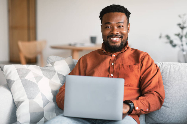 Happy Black Male Using Laptop Computer Smiling To Camera Browsing Internet And Working Online Sitting On Sofa At Home. Freelancer Man Websurfing Indoor. Freelance Concept - Photo, Image