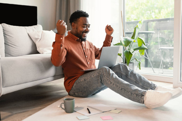 Vreugdevolle zwarte man met behulp van laptop computer werken online en schudden vuisten vieren geweldig nieuws zitten op de vloer thuis. Werk op afstand en leren. Freelance carrièreconcept. Zijaanzicht - Foto, afbeelding