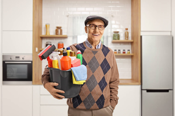 Elderly man holding a bucket with cleaning supplies and standing inside a kitchen - Photo, image