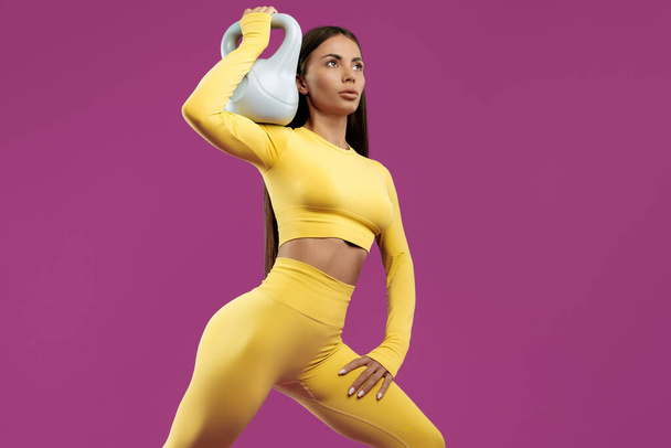 Fit young brunette woman doing exercises with gray kettlebell over purple background - Fotografie, Obrázek