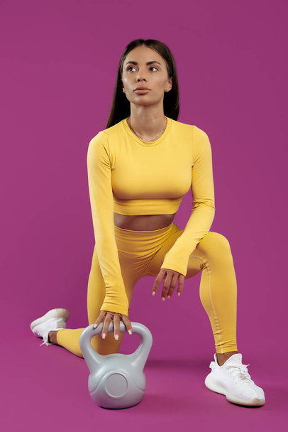 Sporty young brunette woman resting after exercises with gray kettlebell over purple background - Fotó, kép